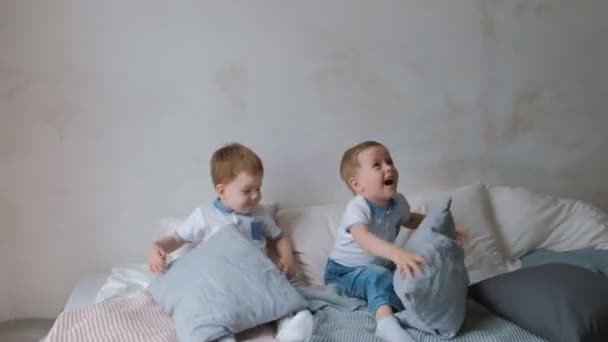 Los niños gemelos están acostados en la cama, tirando almohadas, saltando y riendo . — Vídeos de Stock