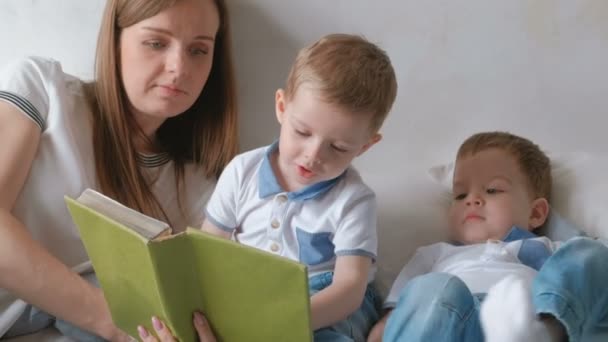 家族ママと双子の兄弟の 2 つの幼児は、ベッドの上に敷設本を読みます。家族の読書の時間. — ストック動画