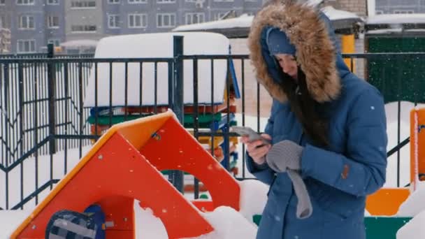 Мама звонит по мобильному телефону, пока ее сын ходит по детской площадке зимой во время снегопада. . — стоковое видео