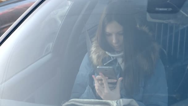ダウン ジャケット、携帯電話を見て青の若いブルネットの女性の車のフロント ガラスを介して表示します。. — ストック動画