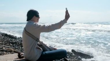 Kadın sonbaharda taş plajda denizin kenarında oturan selfie yapar.