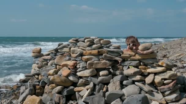 Boy playing, hiding behind a fortress of stones on the sea shore beach. — Stock Video