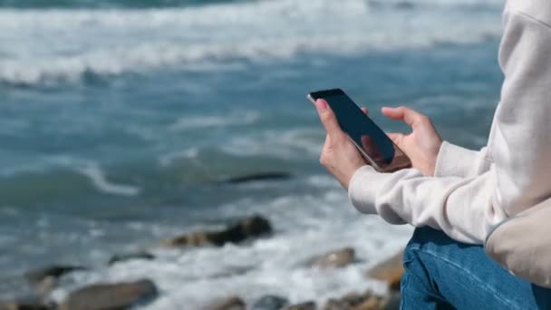 Женщина читает что-то по телефону, сидя на берегу моря осенью. Руки крупным планом . — стоковое видео