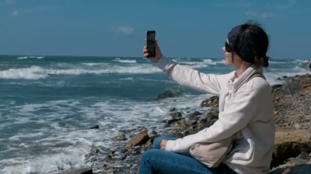 Mujer hace selfie sentado junto al mar en la playa de piedra en otoño . — Vídeo de stock