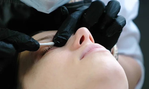 Χέρια cosmetologists τρίβει τα βλέφαρα πελάτες με μια πατσαβούρα βαμβακιού. Botox και μαστίγιο πλαστικοποίηση. Πλάγια όψη. — Φωτογραφία Αρχείου