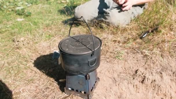 認識できない男はストーブ屋外の芝生に座って森で金属キャンプの炉の鍋料理します。足のクローズ アップ. — ストック動画