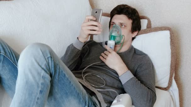 Χρησιμοποιούν νεφελοποιητή και συσκευή εισπνοής για τη θεραπεία. Νεαρός άνδρας εισπνοή μέσω εισπνοής μάσκα ξαπλωμένος στον καναπέ και κουβεντιάζοντας σε κινητό τηλέφωνο. Πλάγια όψη. — Αρχείο Βίντεο