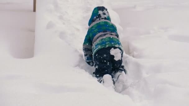 Jongen kruipt op een houten dia omhoog in de sneeuw tijdens de sneeuwval in de tuin van een gebouw met meerdere verdiepingen. — Stockvideo