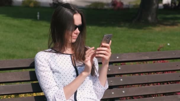 Mujer joven morena escribe un mensaje en el teléfono móvil sentado en el banco en el parque . — Vídeo de stock