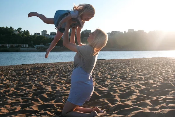 Piękny szczęśliwy Blondynka Mama i Córka, przytulanie i mówiąc, siedząc na plaży o zachodzie słońca. — Zdjęcie stockowe