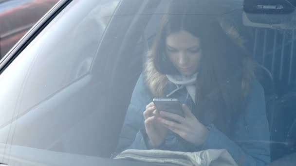Blick durch die Windschutzscheibe des Autos auf junge brünette Frau in blauer Daunenjacke, die auf das Telefon schaut. — Stockvideo