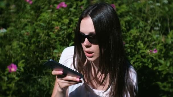 美しいブルネットの女性は、晴れた日に公園で座っている彼女の携帯電話に音声メッセージを記録します。. — ストック動画