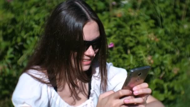 Жінка друкує повідомлення на мобільному телефоні, сидячи в парку в сонячний день, спілкуючись . — стокове відео