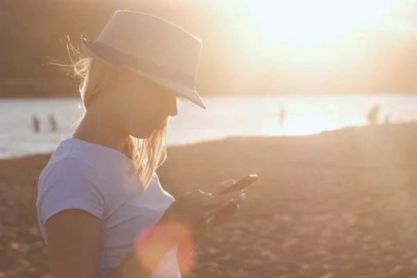 Красивая блондинка в шляпе печатает сообщение на мобильном телефоне на пляже на закате . — стоковое фото