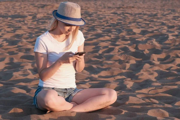 Красивая блондинка в шляпе печатает сообщение на мобильном телефоне на пляже на закате . — стоковое фото