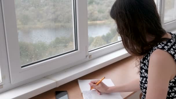 Женщина пишет письмо в блокноте, сидя дома на балконе. Вид на реку. Дождь за окном . — стоковое видео