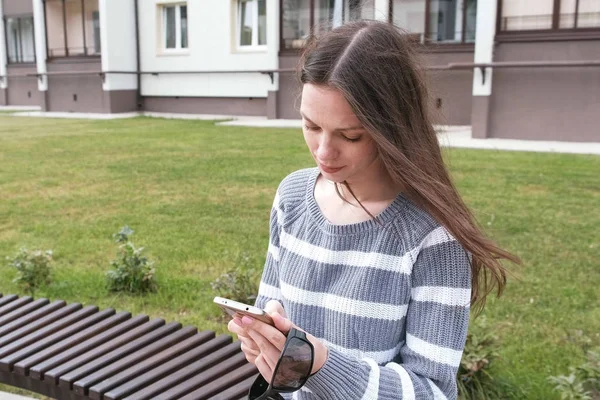 Attraktive brünette Frau plaudert und tippt Nachricht auf Handy und lächelt auf der Bank sitzend. — Stockfoto