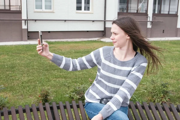 Linda morena está tentando tirar uma selfie em seu telefone celular ao vento enquanto se senta no banco . — Fotografia de Stock