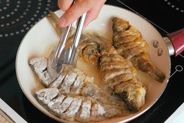 O homem fritou o peixe no óleo em uma panela. Mãos de homem close-up vira peixe com pinças culinárias . — Fotografia de Stock