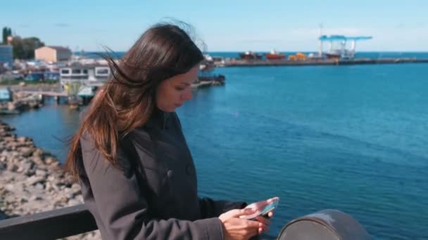 Mujer morena escribe un mensaje en un teléfono móvil de pie en el paseo marítimo junto al mar en un día soleado de otoño . — Vídeo de stock