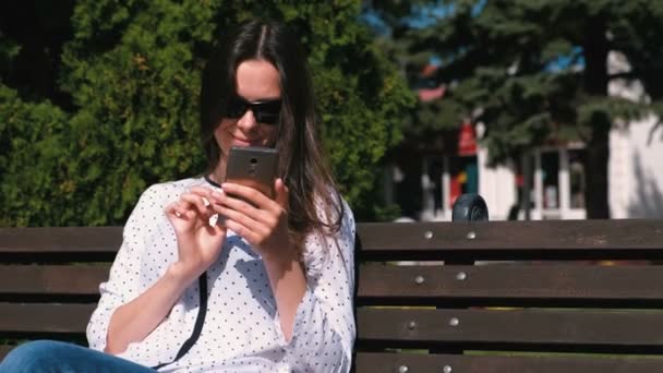 Jonge vrouw brunette typt een bericht op mobiele telefoon zittend op het bankje in park. — Stockvideo