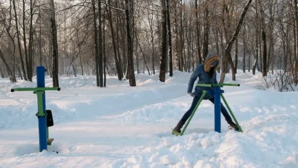 Γυναίκα με μπλε χρώμα κάτω σακάκι με κουκούλα κάνει ασκήσεις ποδιών σε προσομοιωτή σε ένα χειμερινό πάρκο μπροστινή όψη — Αρχείο Βίντεο