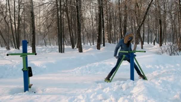 Vrouw in blauw donsjack met kap doet benen oefeningen op de simulator in een winter Park vooraanzicht — Stockvideo