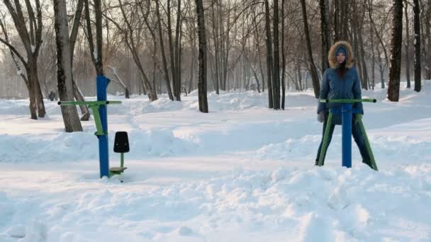 Γυναίκα με μπλε σακάκι κάνει πόδια ασκήσεις σε προσομοιωτή σε ένα χειμερινό πάρκο μπροστά αριστερά Δες στο βίντεο. — Αρχείο Βίντεο