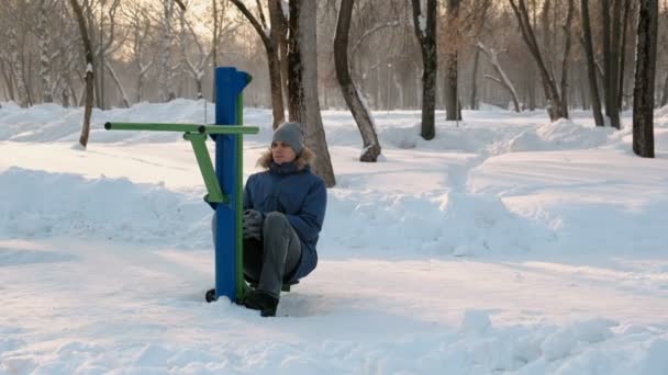 Чоловік в синій куртці з капюшоном робить вправи на ногах на симуляторі в зимовому парку. Вид спереду . — стокове відео