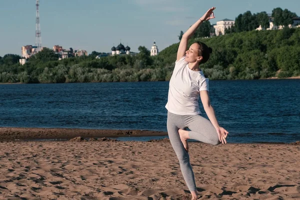 市内の川のビーチでヨガのストレッチの女性。美しいシティー ビュー。Vrikshasana ポーズ. — ストック写真