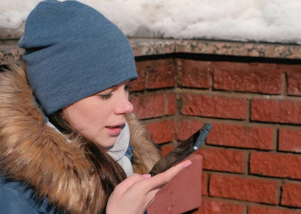Woman is speaking audio message on mobile phone sitting in winter park. Closeup face side view.