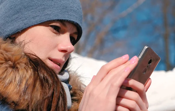 Mujer está navegando páginas de Internet en el teléfono móvil sentado en el parque de invierno. Cara de primer plano . — Foto de Stock