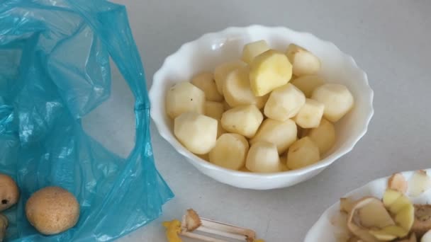 Batatas pequenas em prato, casca e descascador na mesa da cozinha. Batata descascada no saco azul de plástico . — Vídeo de Stock