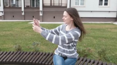 Güzel esmer bankta otururken rüzgarda cep telefonunda bir selfie almaya çalışıyor.