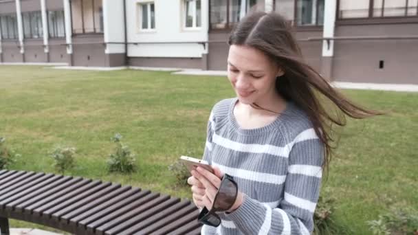 Attraktiv brunett kvinna chatta och skriva meddelande på mobiltelefon och leende sitter på bänken. — Stockvideo