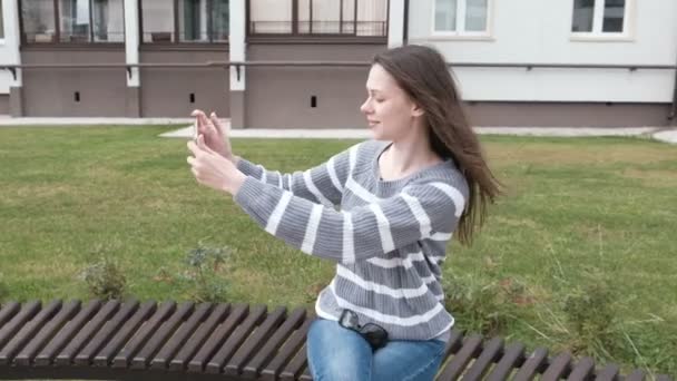 Hermosa morena está tratando de tomar una selfie en su teléfono móvil en el viento mientras está sentada en el banco . — Vídeos de Stock