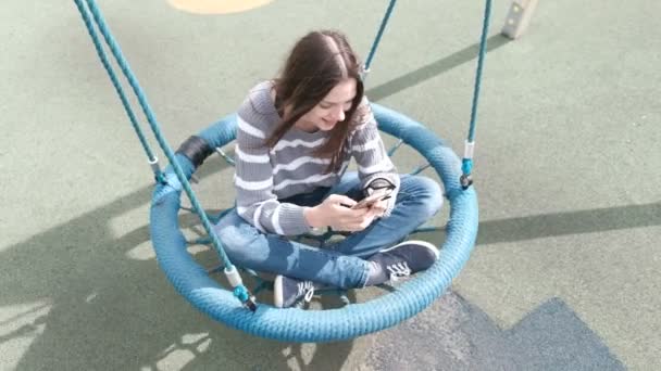 携帯電話でメッセージを入力して、美しいブルネットの少女は空の水かきのあるスイングでスイングします。. — ストック動画