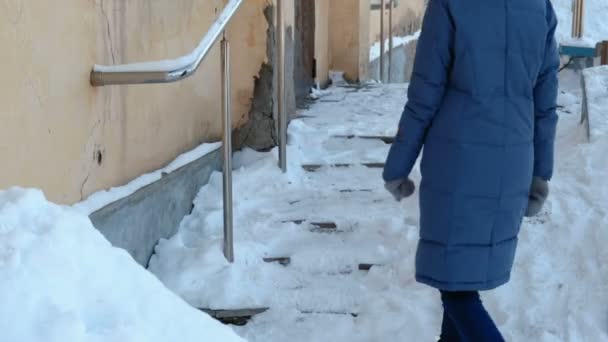 Slippery stairs. Unrecognizable woman in a blue down jacket walking up a snowy staircase in street. — Stock Video