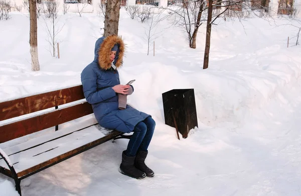 Mulher está sentada no banco e navegando telefone celular no parque de inverno na cidade durante o dia . — Fotografia de Stock