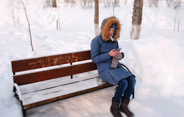 여 자가 벤치에 앉아서 화창한 날에 겨울 공원 도시에서 휴대 전화를 탐색. — 스톡 사진
