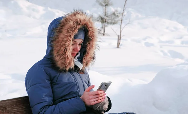 Mulher está sentada no banco e navegando telefone celular no parque de inverno na cidade em dia ensolarado . — Fotografia de Stock