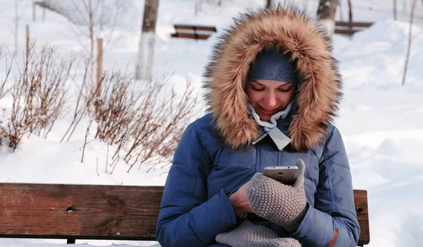 Улыбающаяся женщина сидит на скамейке и просматривает мобильный телефон в зимнем парке в городе в солнечный день . — стоковое фото