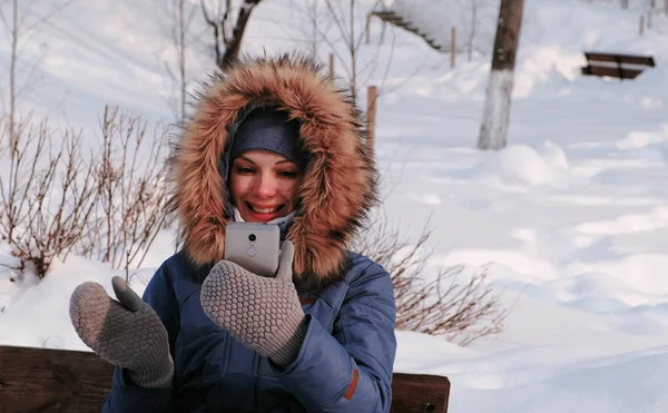 Jovem mulher bonita feliz chamada vídeo conversando no parque de inverno na cidade em dia nevado com queda de neve . — Fotografia de Stock