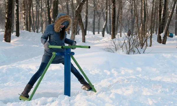 Mulher de casaco azul para baixo com capuz está fazendo exercícios de pernas no simulador em um parque de inverno Vista frontal — Fotografia de Stock