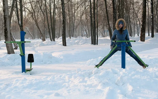 Mulher de casaco azul para baixo com capuz está fazendo exercícios de pernas no simulador em um parque de inverno Vista frontal — Fotografia de Stock