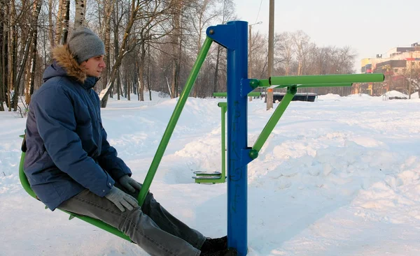 남자 후드 재킷 아래로 파란색 겨울 공원에서 시뮬레이터에 다리 운동을 하 고 있다. 측면 보기. — 스톡 사진