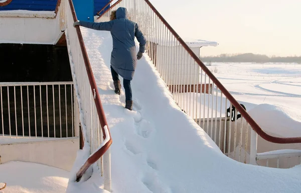 ブルー ジャケットで認識できない女性は、雪に覆われた階段を登っていきます。背面図. — ストック写真