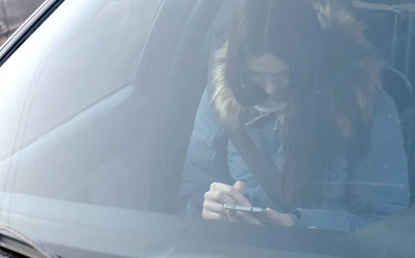 Vue à travers le pare-brise de la voiture sur la jeune femme brune en duvet bleu regardant le téléphone . — Photo