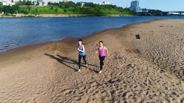 İki kadın nehir kumlu plaj boyunca gün batımında koşu. Güzel şehir manzaralı. — Stok video