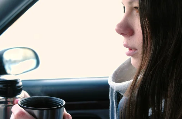Jovem morena bebe chá de uma garrafa térmica sentada em um carro no inverno . — Fotografia de Stock
